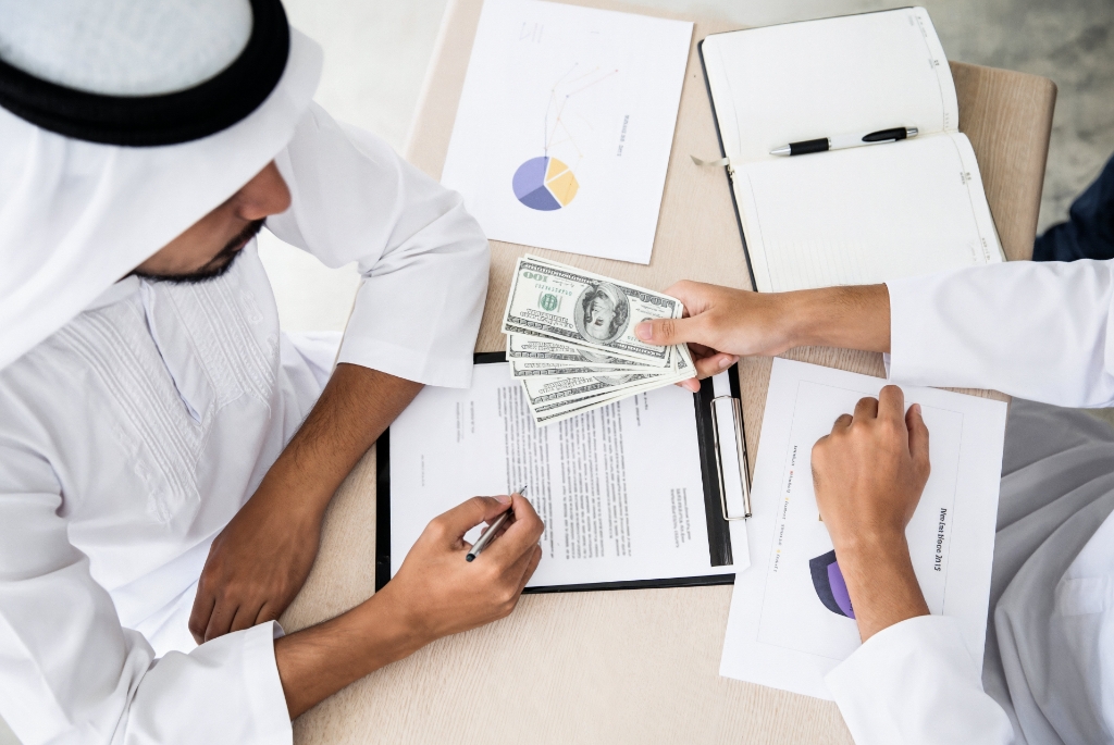 A Person Handing Money to an Arab Individual While Both are Seated at a Table